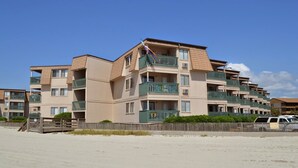 Un immeuble résidentiel à plusieurs étages, doté de balcons, avec un rez-de-chaussée dont le sol est recouvert de sable, et un ciel d’un bleu limpide.