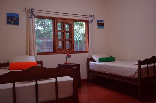 A room with two wooden beds, a window with curtains, and a small table with a lamp.