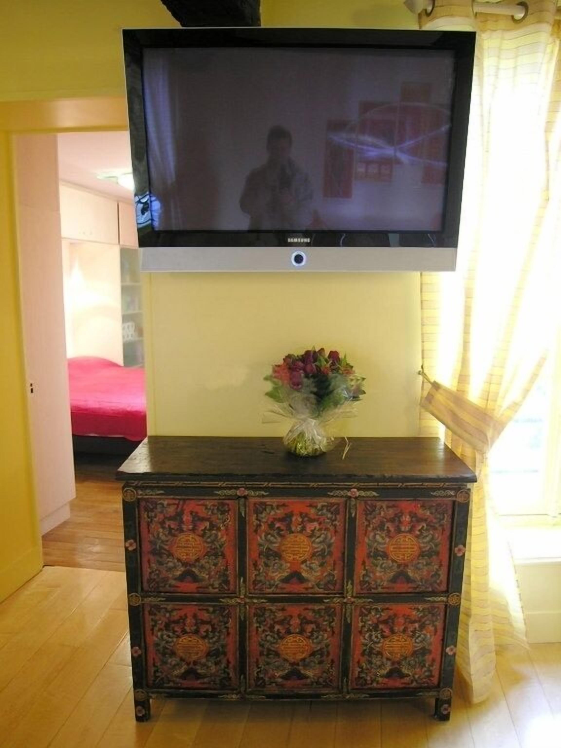 A room with a television mounted on the wall, a floral arrangement on a wooden cabinet, and a bed with a red cover.