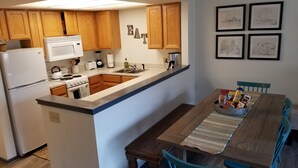 Een keuken met houten kastjes, een witte koelkast, een fornuis, een magnetron en een spoelbak. Er staat een eettafel met een placemat en een schaal met lekkernijen.