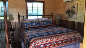 A wooden cabin bedroom with a bed, two bedside lamps, a window, and an air conditioning unit. - Crooked Creek Ranch Cabin 2 (Hamilton)