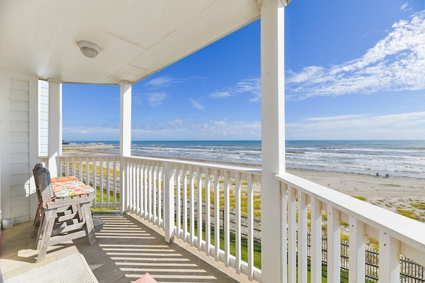 A covered porch with a view of the ocean, a wooden chair, and a table with a colorful cushion.