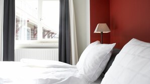 A hotel room with a bed, a window with curtains, a red wall, and a lamp. - Hotel De Prins (Sittard)