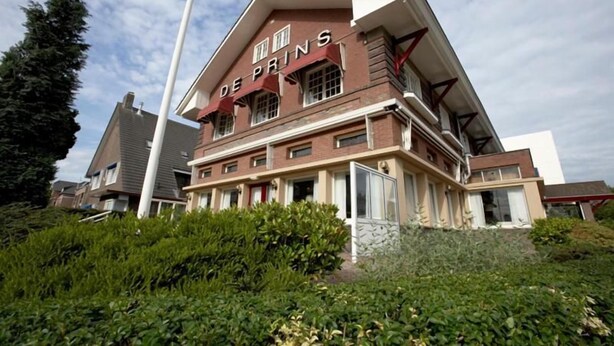 Un edificio de dos pisos con el nombre 'DE PRINS' en la fachada, rodeado de follaje.