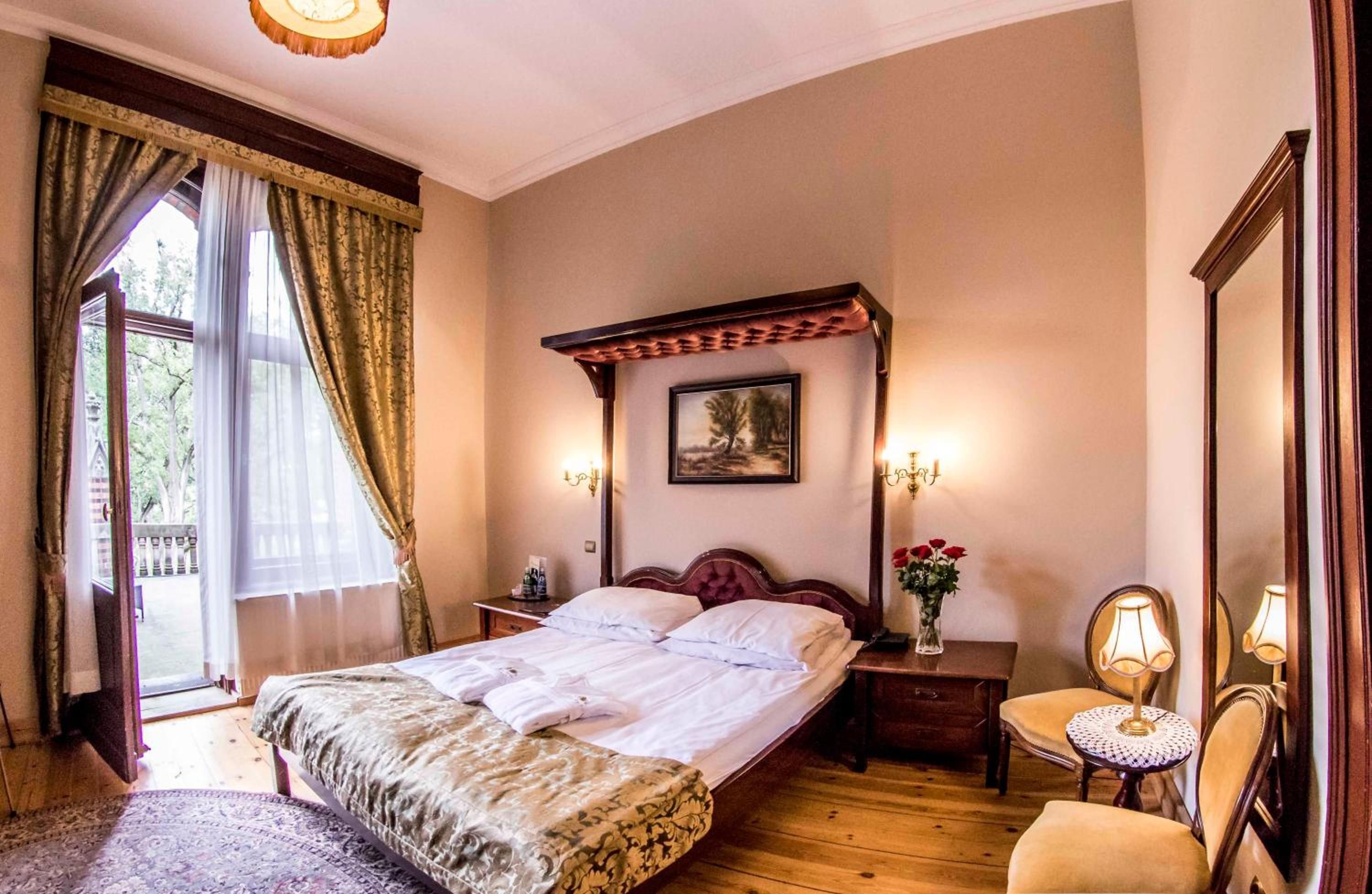 A hotel room with a large bed, a wooden headboard, a nightstand with a lamp, a small table with a vase of flowers, and a window with curtains.