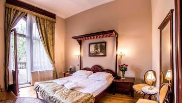 A hotel room with a large bed, a wooden headboard, a nightstand with a lamp, a small table with a vase of flowers, and a window with curtains.