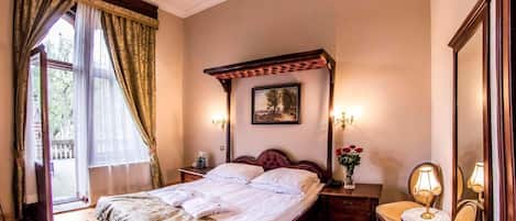 A hotel room with a large bed, a wooden headboard, a nightstand with a lamp, a small table with a vase of flowers, and a window with curtains.