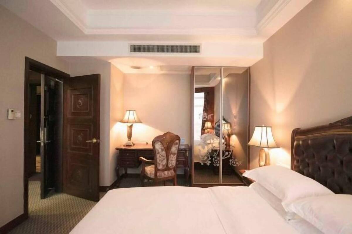 Una habitación de hotel con cama, sillón, escritorio, lámpara, espejo y puerta.