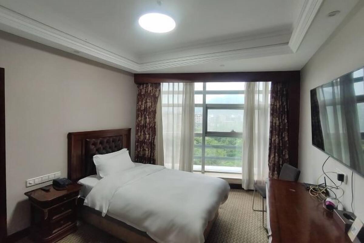 Una habitación de hotel con cama, escritorio, televisor y ventana con cortinas.