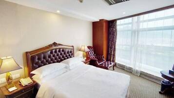 A hotel room with a large bed, a wooden headboard, a nightstand with a lamp, a chair, and a large window with a view.