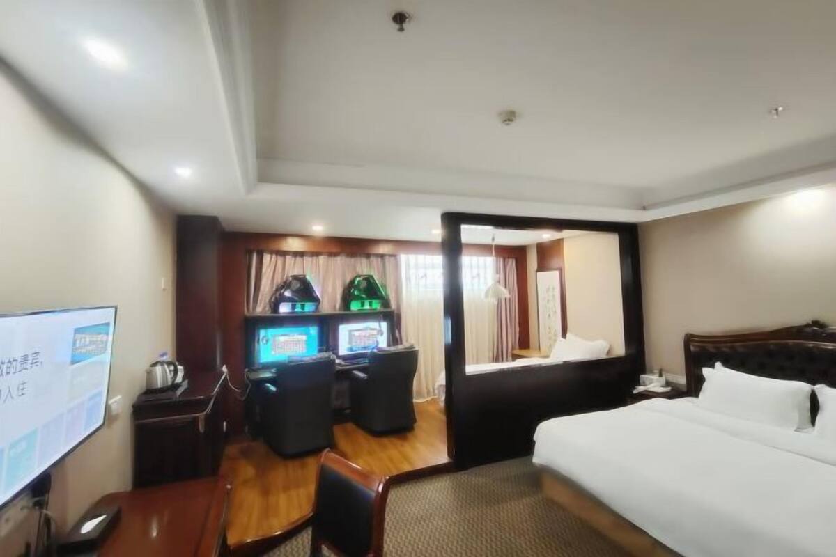 Habitación de hotel con cama, escritorio con computadora, dos sillas gamer, televisor y ventana con cortinas.