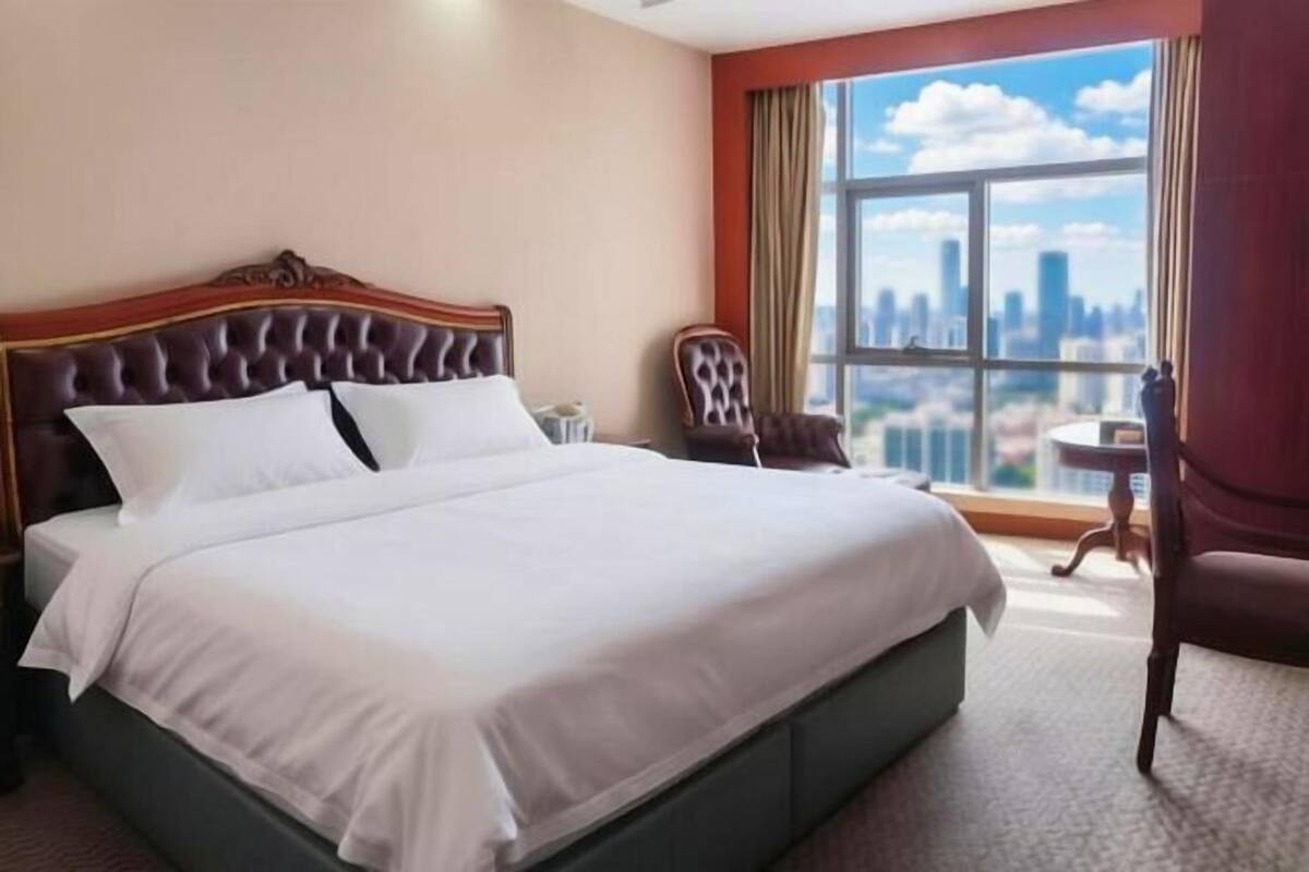 Habitación de hotel con una cama grande, una silla, una mesita y vistas a un paisaje urbano a través de un amplio ventanal.