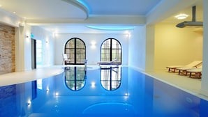Una piscina interior con paredes de vidrio transparente, una pared decorativa de ladrillo y sillones de descanso.