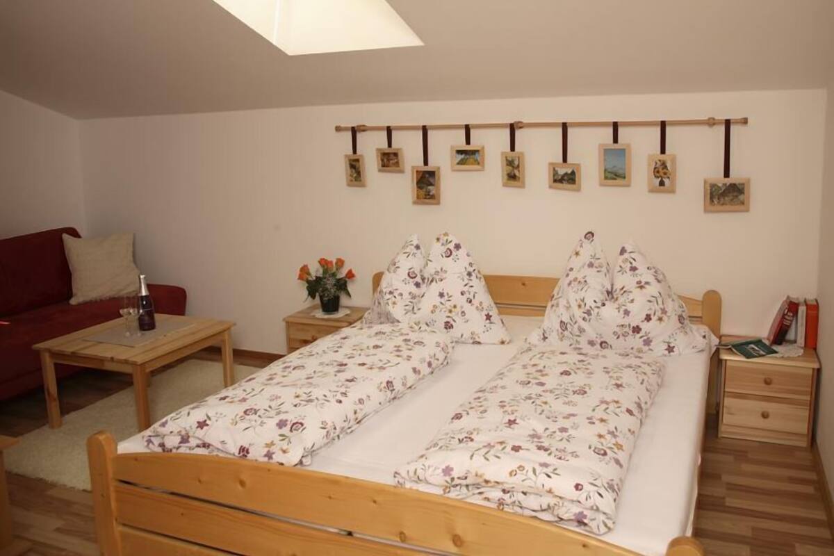 A bedroom with a bed, a bedside table, a nightstand, a red sofa, a small table with a bottle and flowers, and a skylight.
