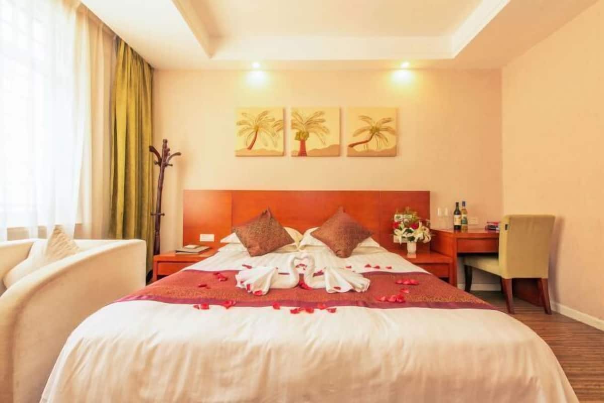 A hotel room with a bed, pillows, a nightstand, a chair, a desk, and a wall with three palm tree paintings.