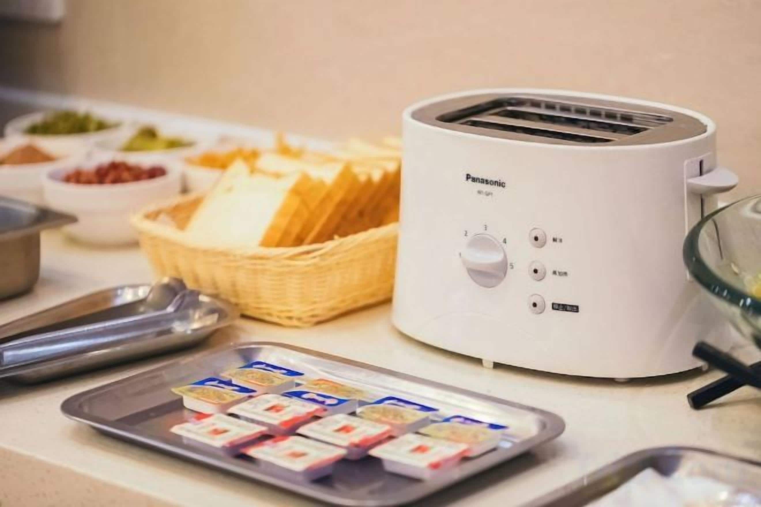 Un tostador Panasonic sobre una encimera de cocina con una cesta de pan, una bandeja de alimentos envasados y una cuchara de metal.