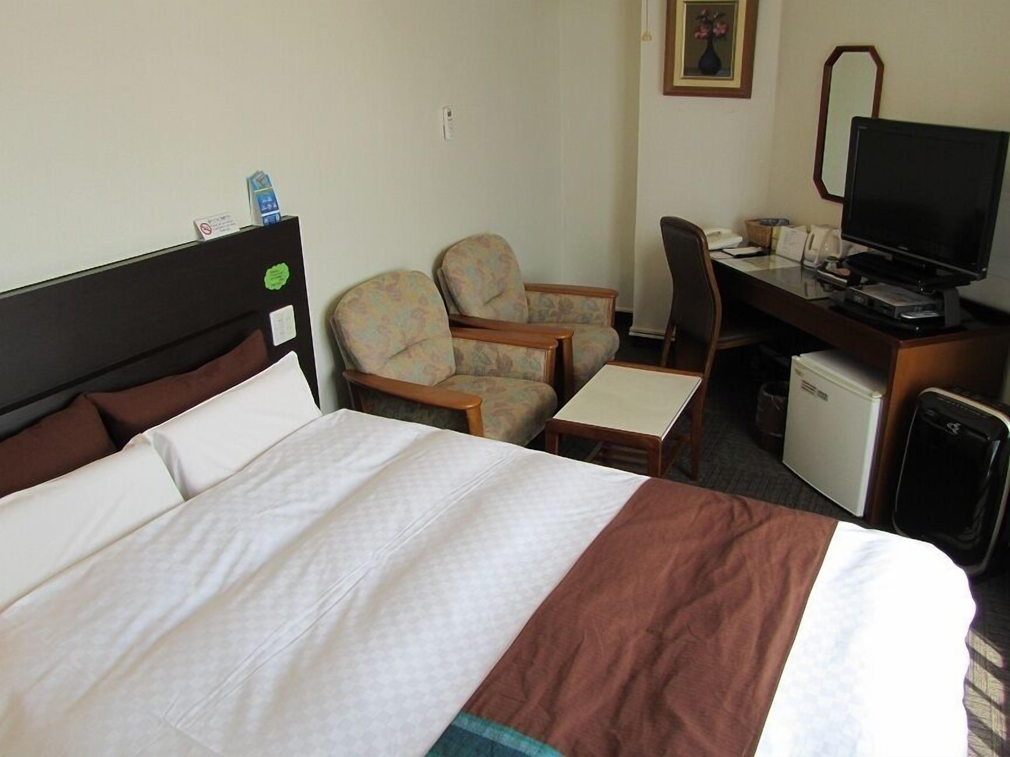 A hotel room with a bed, two armchairs, a desk with a computer, a television, and a small refrigerator.