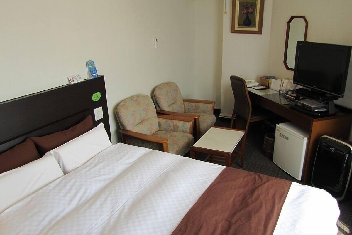 A hotel room with a bed, two armchairs, a desk with a computer, a television, and a small refrigerator.