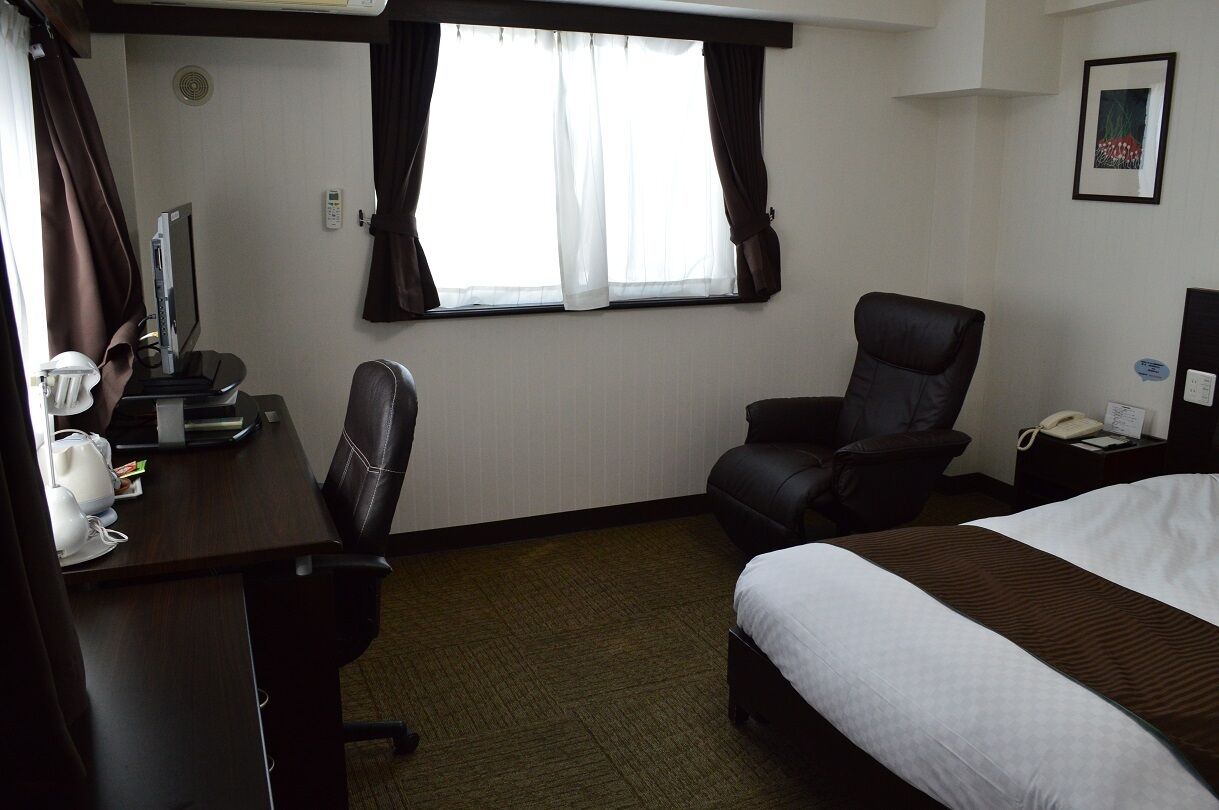 A hotel room with a bed, a desk with a computer, a chair, a window with curtains, and a wall-mounted phone.
