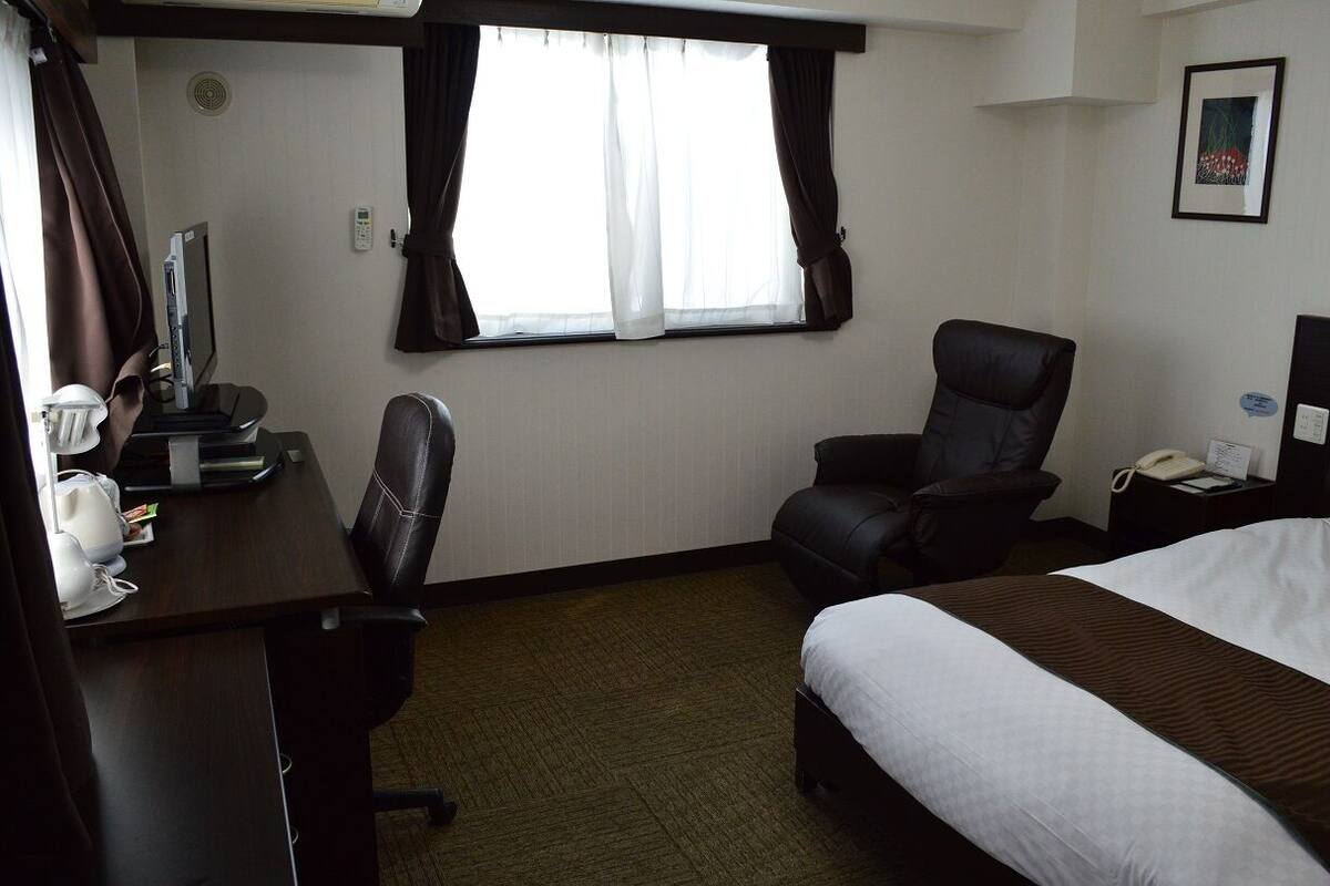 A hotel room with a bed, a desk with a computer, a chair, a window with curtains, and a wall-mounted phone.