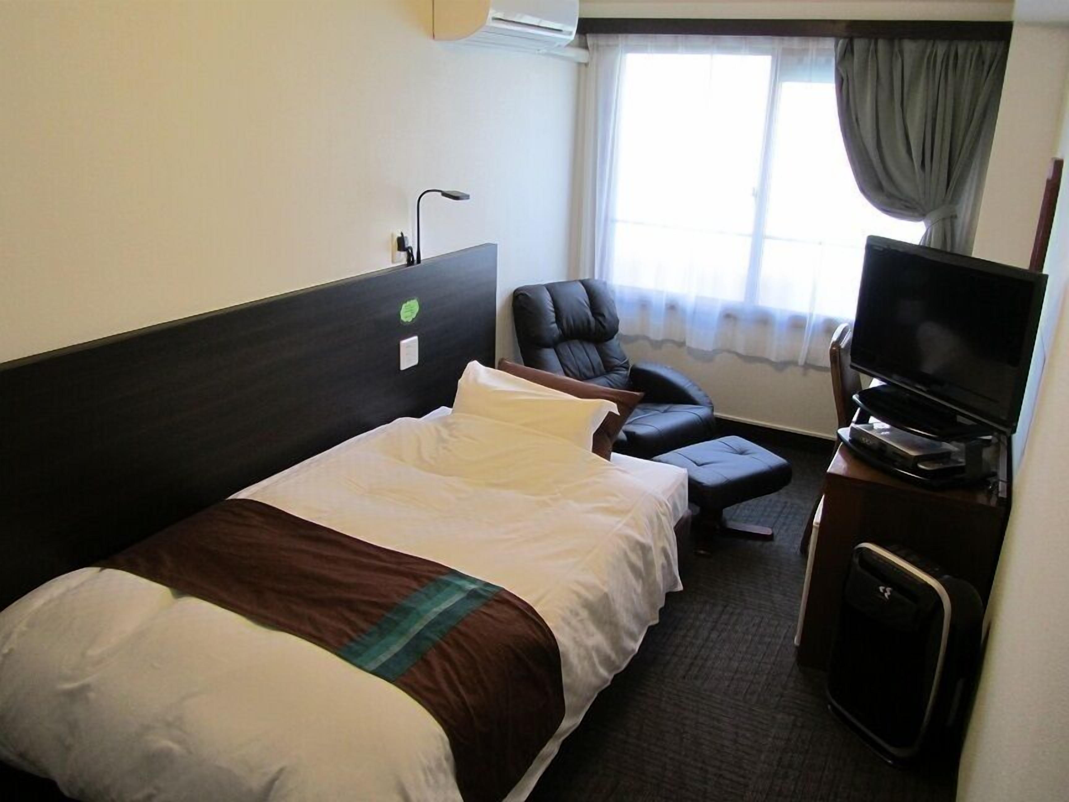 A hotel room with a bed, a chair, a television, and a window with curtains.