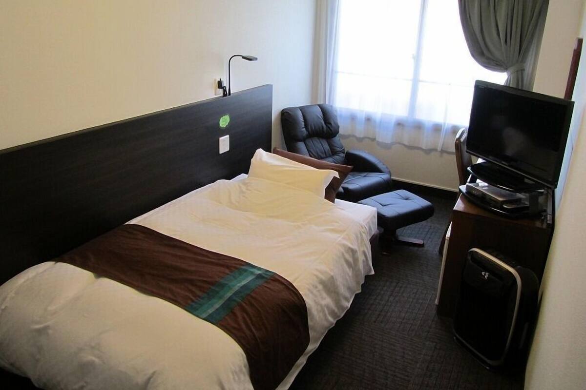 A hotel room with a bed, a chair, a television, and a window with curtains.