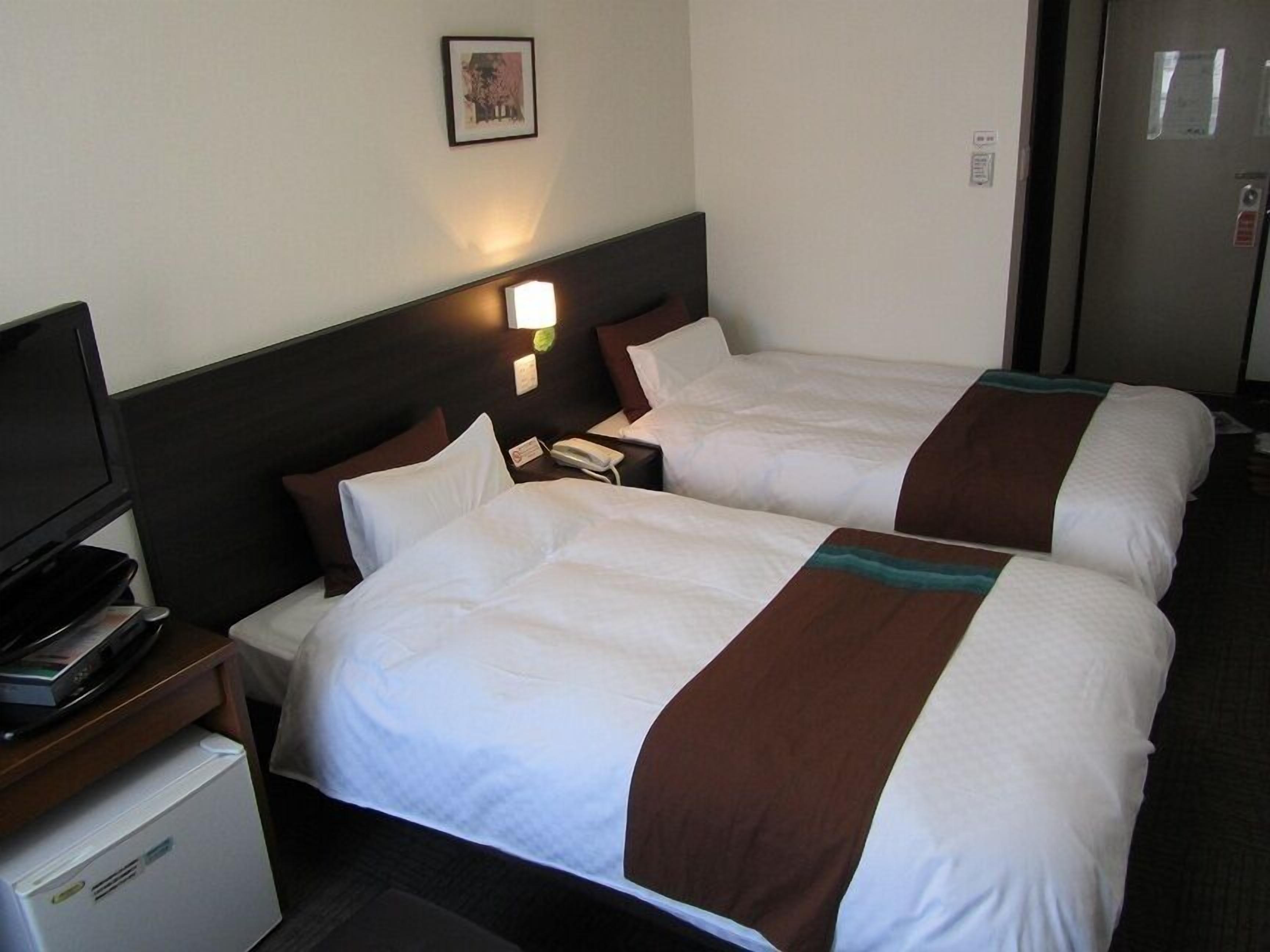 A hotel room with two beds, a TV, a refrigerator, and a wall-mounted light.