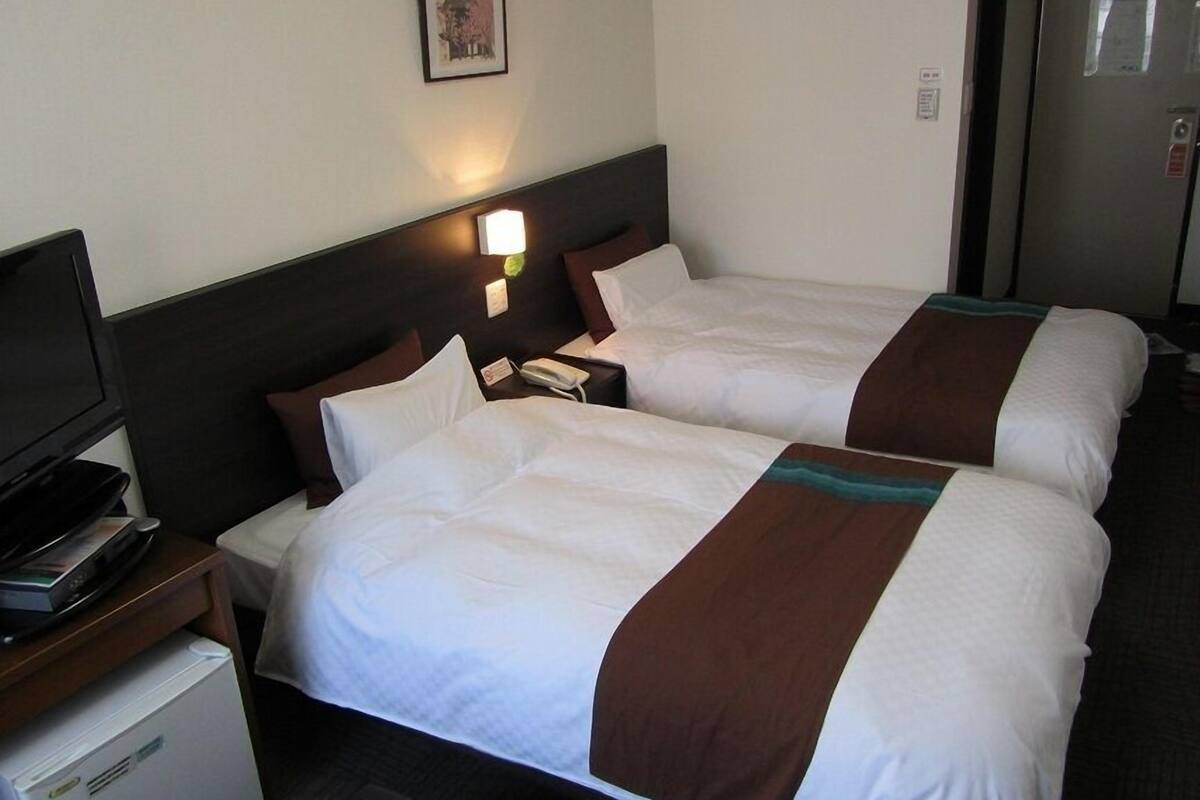 A hotel room with two beds, a TV, a refrigerator, and a wall-mounted light.