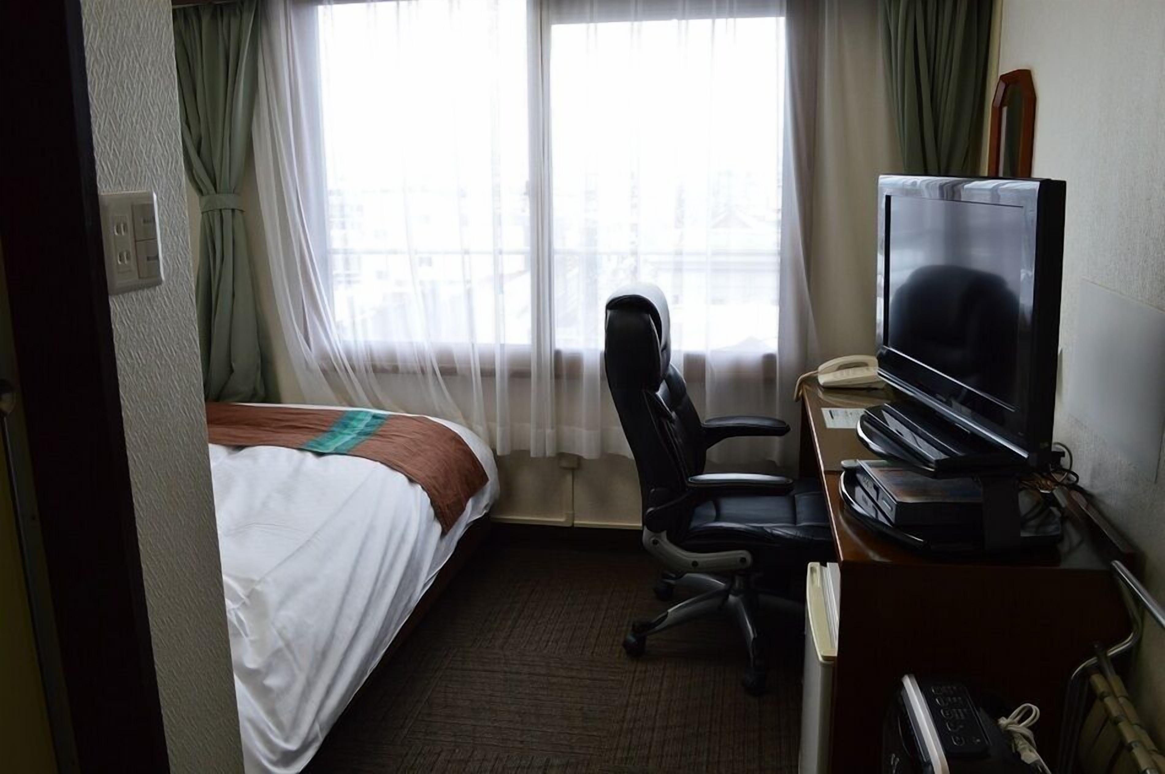 A hotel room with a bed, a desk with a television, a chair, and a window with curtains.