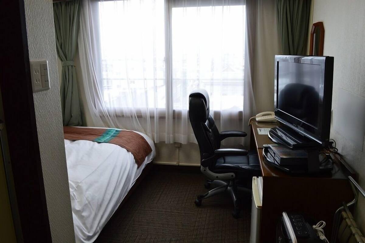 A hotel room with a bed, a desk with a television, a chair, and a window with curtains.