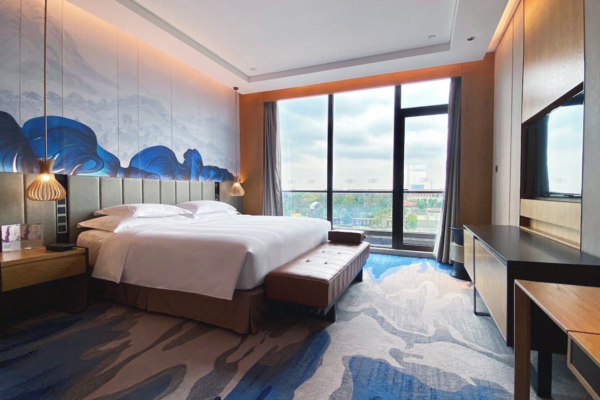 Ein Hotelzimmer mit einem großen Bett, einem Schreibtisch, einem Fernseher und Blick auf die Stadt durch große Fenster.