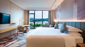 Une chambre d’hôtel avec un grand lit, un bureau, une chaise, une télévision et une vue sur un lac et le paysage urbain.