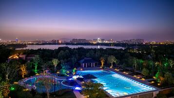 A nighttime view of a swimming pool area with surrounding trees and a distant city skyline.