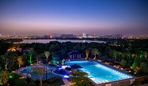 Vista nocturna de una zona de piscina con árboles circundantes y un horizonte urbano distante.