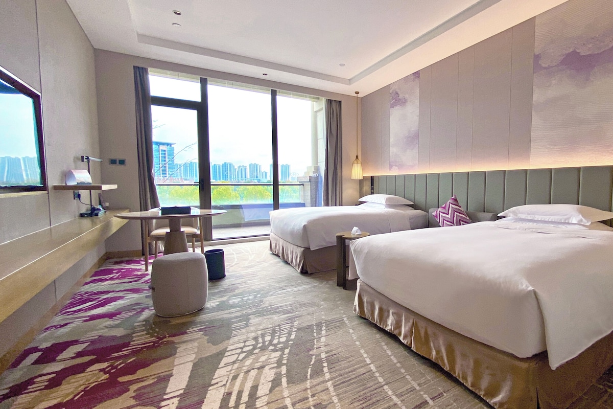 Habitación de hotel con dos camas, un escritorio y un ventanal grande con vistas a la ciudad.