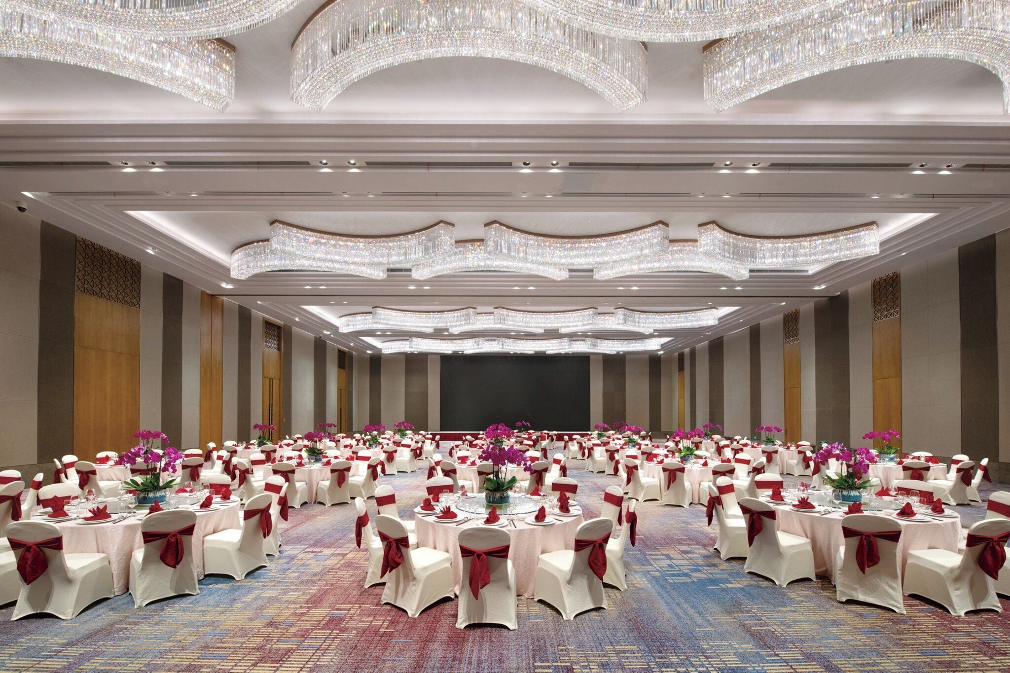 Une grande salle de banquet avec des tables rondes, des chaises blanches et des rubans rouges.
