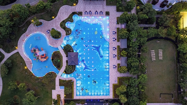 An aerial view of a swimming pool complex with multiple pools, seating areas, and surrounding greenery.
