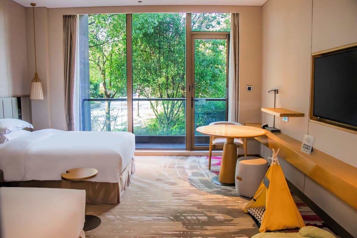 A hotel room with two beds, a small round table, a yellow tent, and a large window with a view of trees.