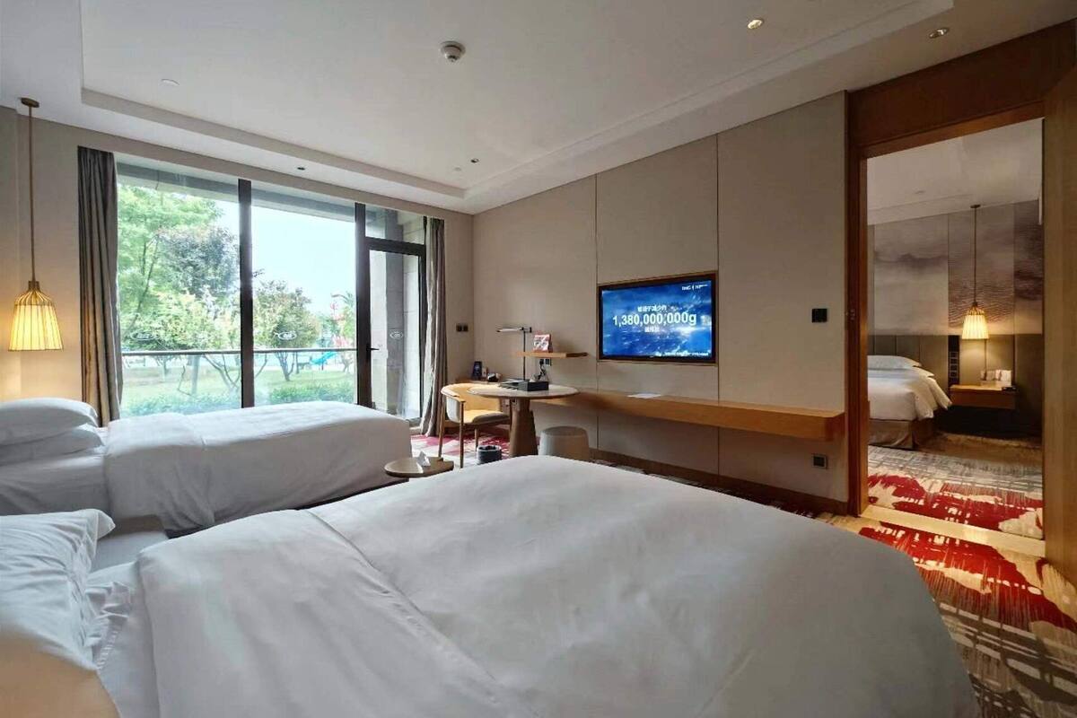 Ein Hotelzimmer mit zwei Betten, einem Schreibtisch, einem Fernseher und einem großen Fenster mit Blick auf Grünflächen.