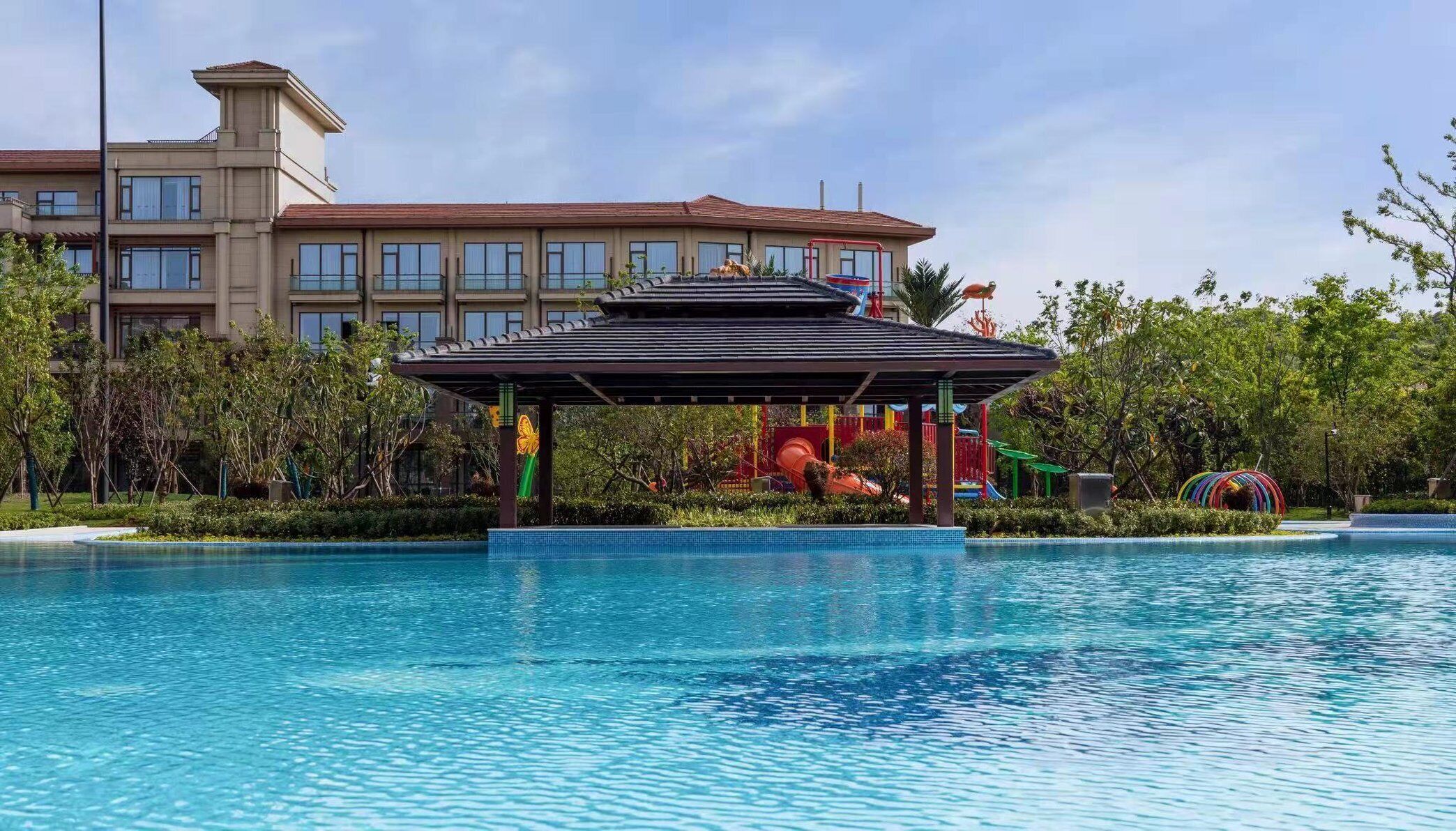 Una piscina con un pabellón, una zona de juegos para niños y un edificio de hotel al fondo.