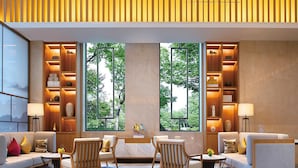 Un salon moderne avec un canapé, des fauteuils et une table basse. De grandes fenêtres offrent une vue sur les arbres. Des étagères ornées d’objets décoratifs sont disposées de part et d’autre des fenêtres.