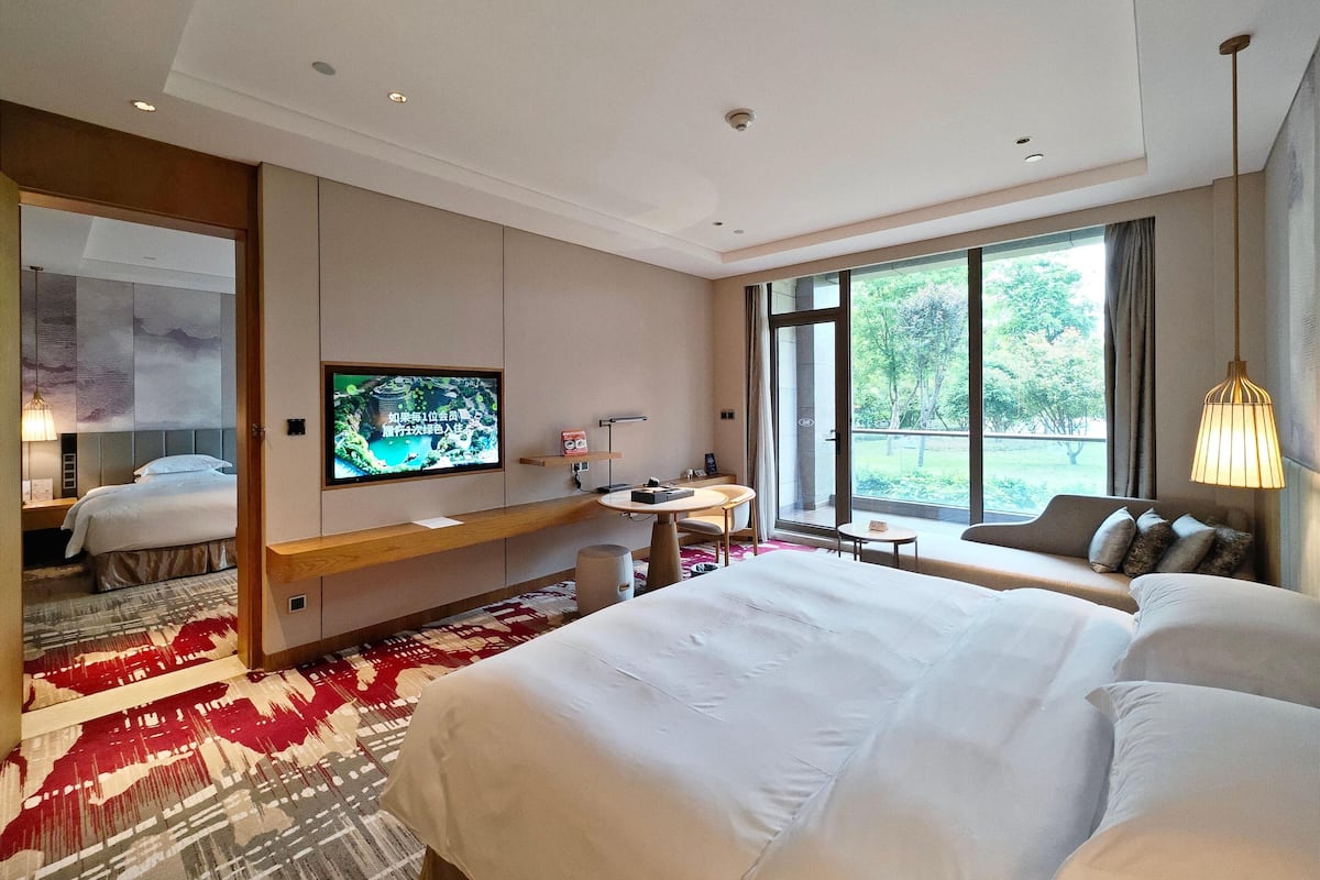 Ein Hotelzimmer mit einem großen Bett, einem an der Wand montierten Fernseher, einem Schreibtisch mit Stuhl, einem kleinen Tisch und einem Sofa.