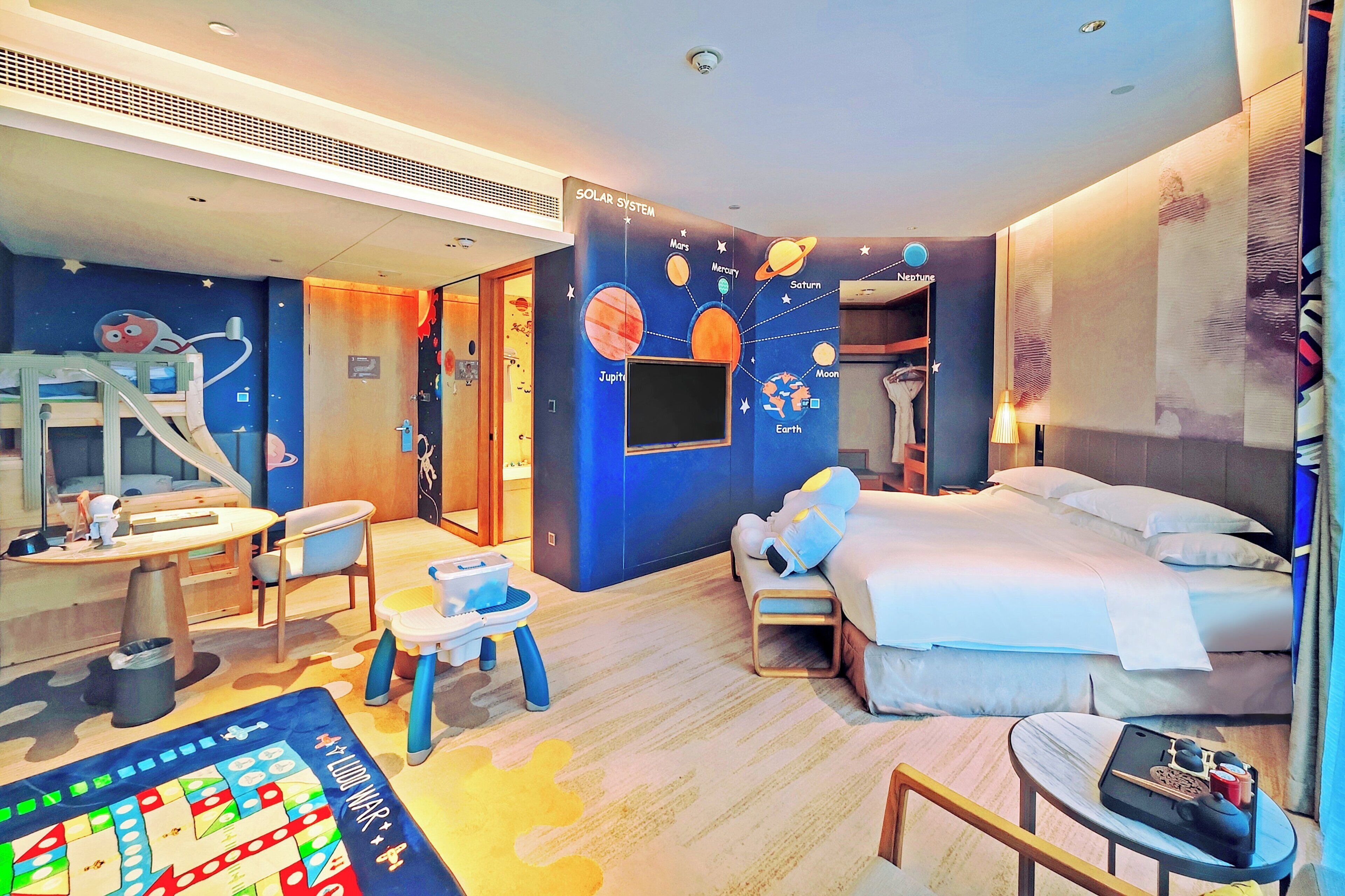 A hotel room with a bed, a desk, a chair, a small table, a child’s play area, and a wall with a solar system mural.