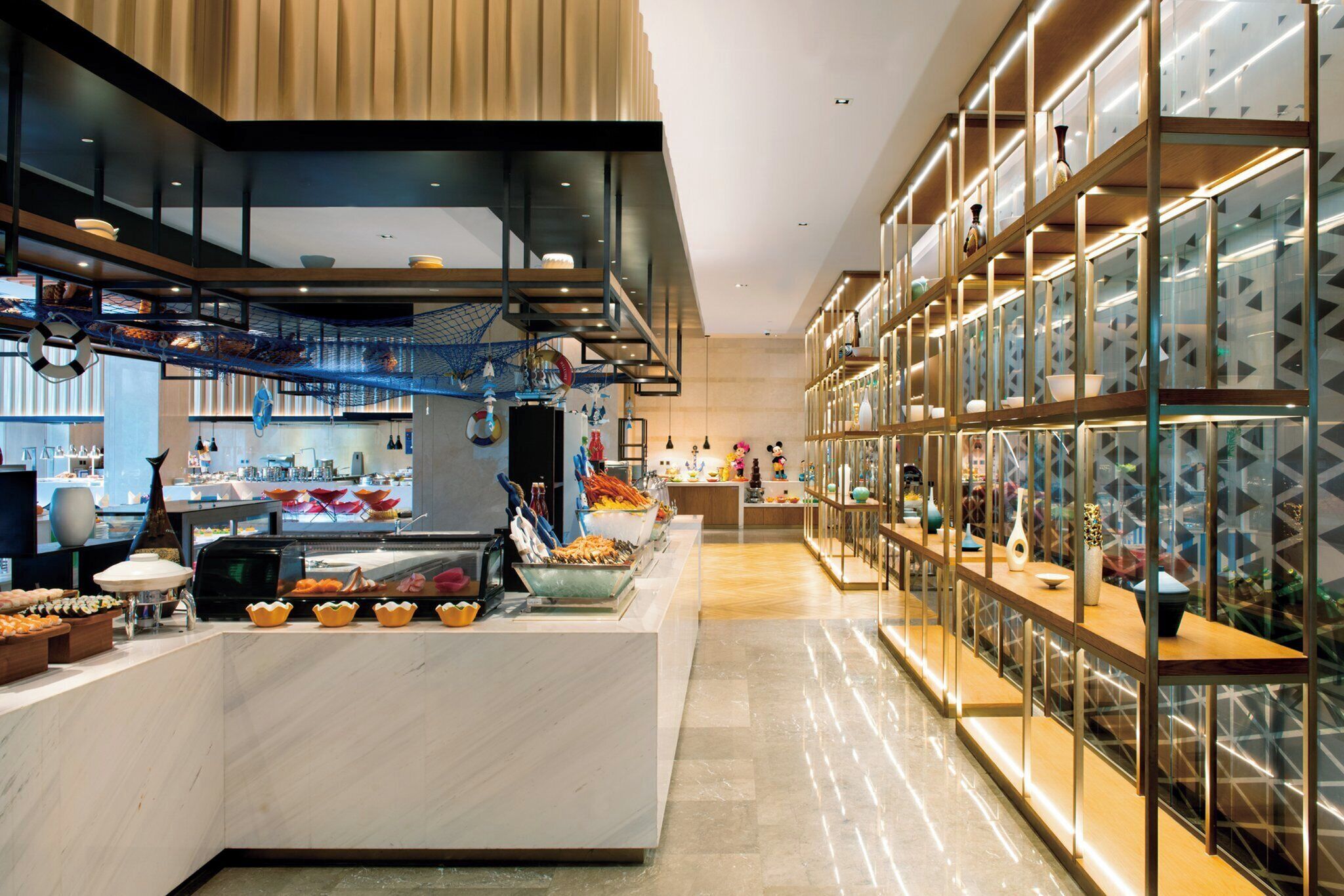 A modern hotel lobby with a buffet area, a seating area with tables, and a decorative wall with patterned glass panels.
