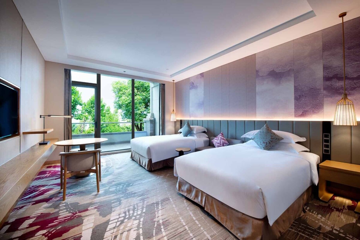 A modern hotel room with two beds, a small table, a TV, and a large window offering a view of greenery.