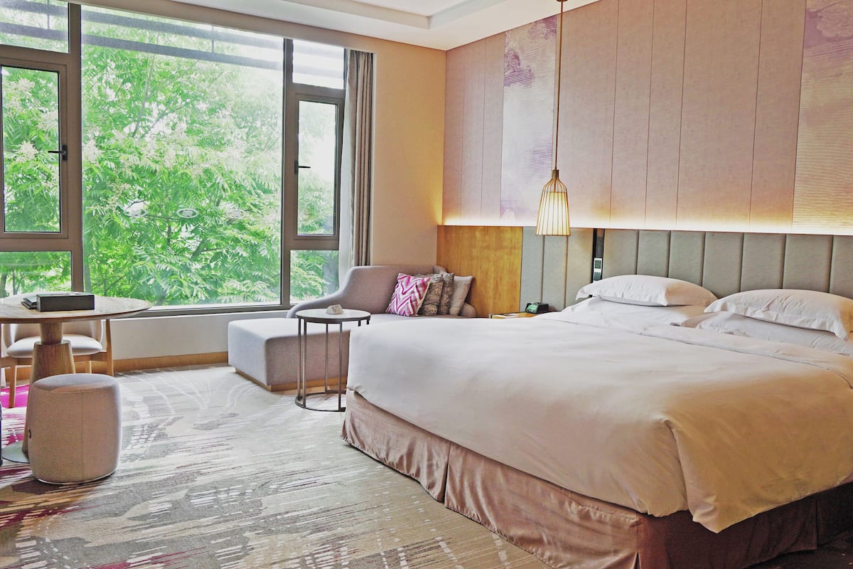 Ein Hotelzimmer mit einem großen Bett, einem kleinen runden Tisch, einem Stuhl und Blick auf Grün hinter dem Fenster.