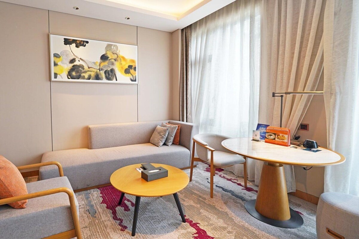 A modern hotel room with a sofa, armchair, round table, and a painting on the wall.