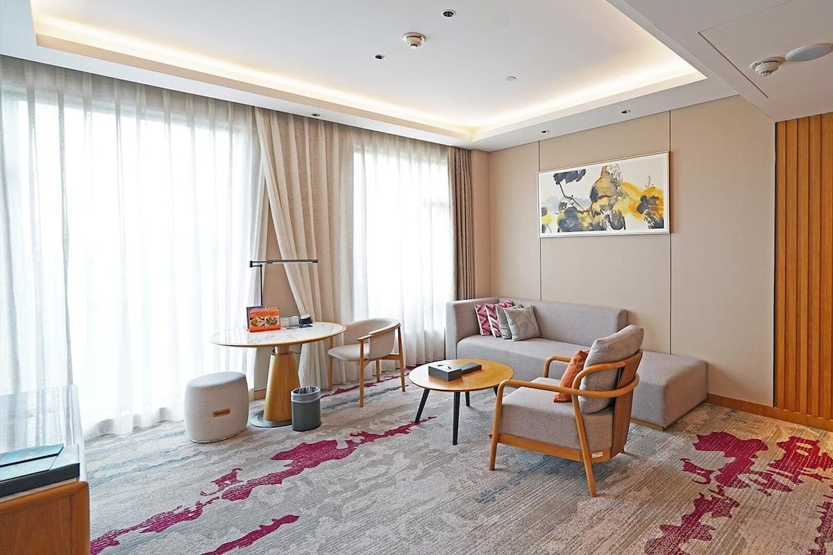 Ein modernes Hotelzimmer mit einer Sitzecke, einem kleinen runden Tisch, einem Sessel und einem großen Fenster mit Vorhängen.