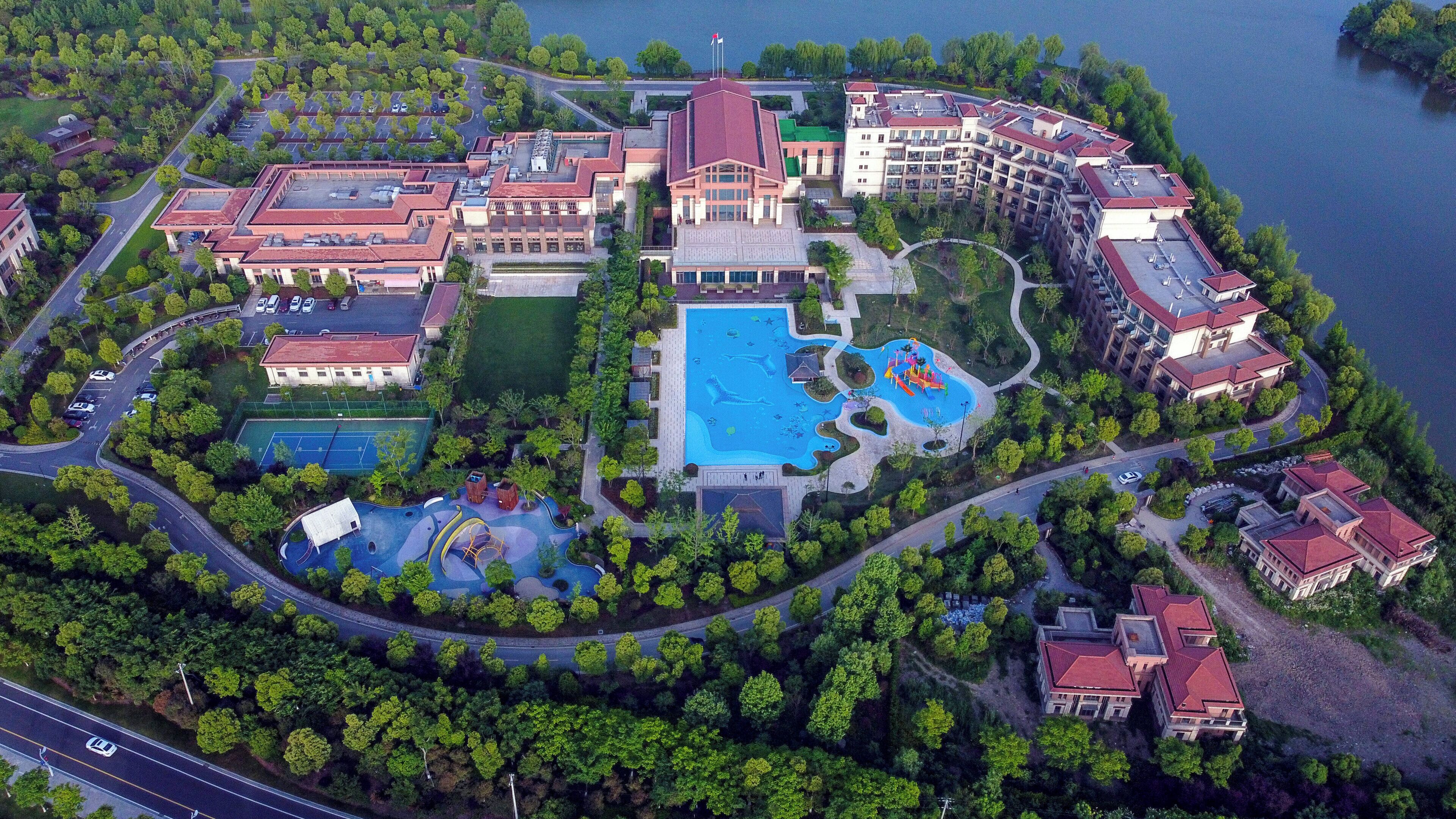 An aerial view of a resort with multiple buildings, a central swimming pool, and surrounding greenery.