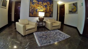 Un vestíbulo con dos sillones beige, una alfombra estampada, un cuadro abstracto grande y una mesita con una lámpara.